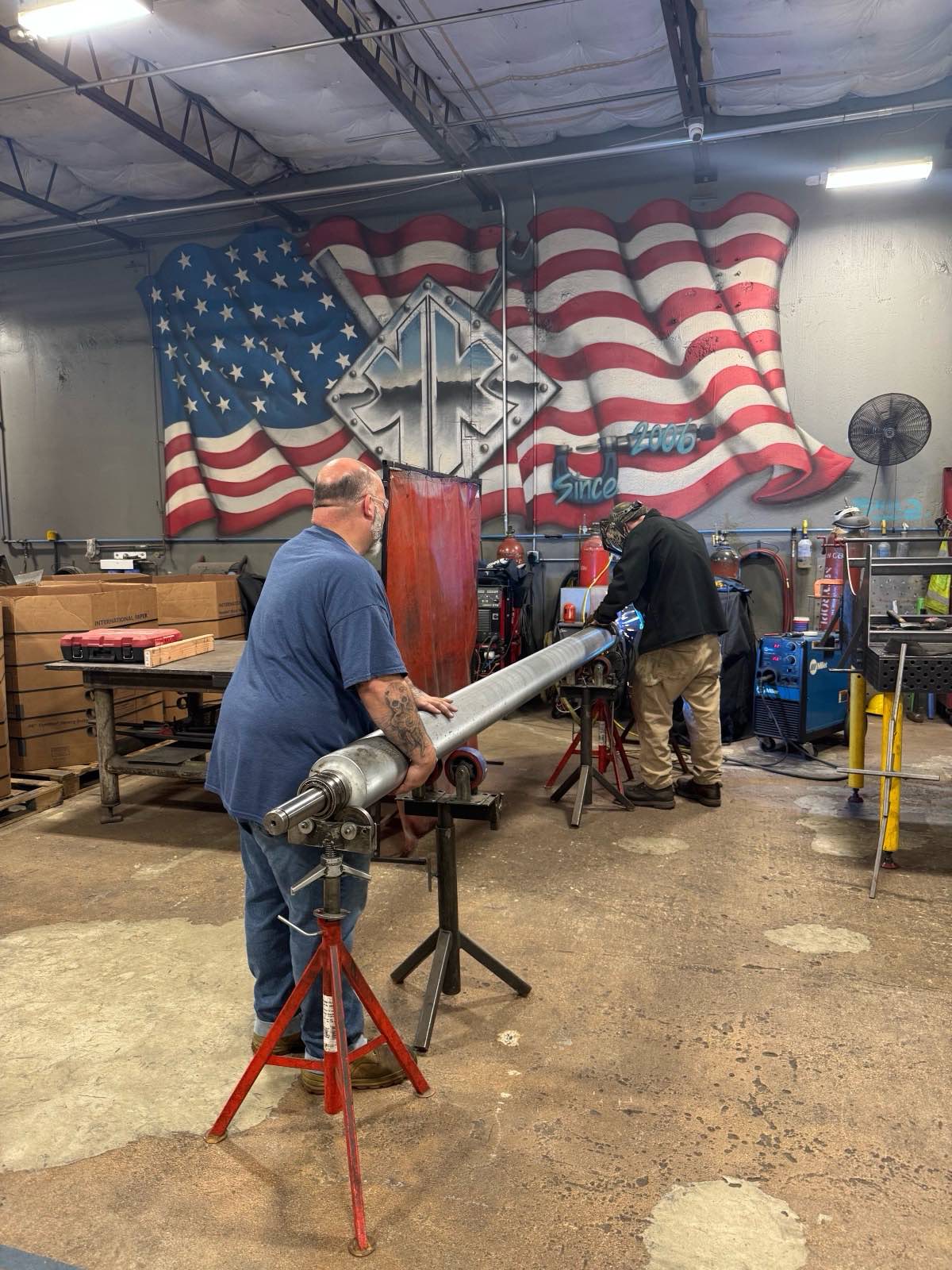 Kimbrell team members working a long shaft on roller stands in front of the shop's American-flag mural